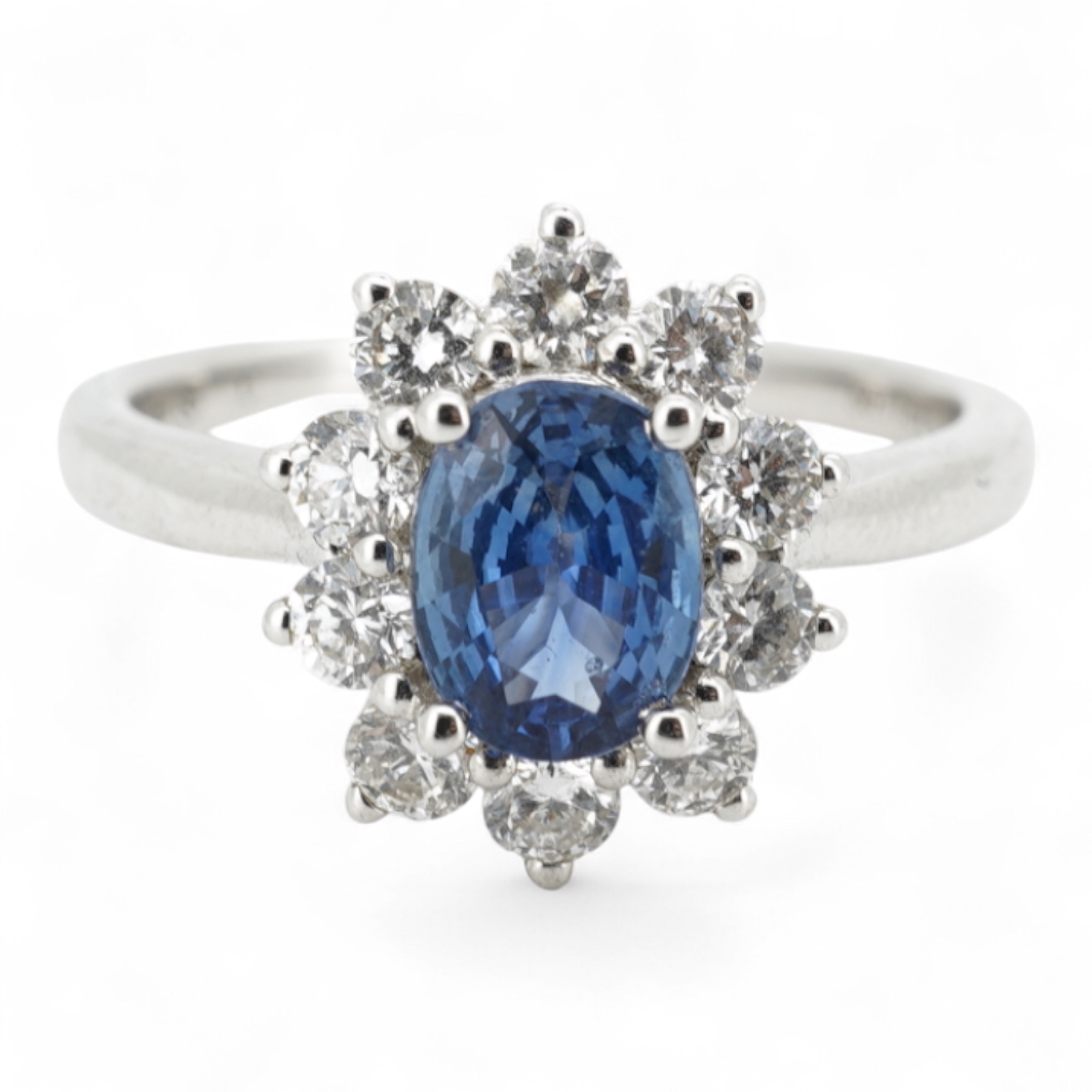 A sapphire and diamond cluster ring, claw-set with an oval sapphire weighing approximately 0.77 carats, within a border of brilliant-cut diamonds totalling approximately 0.50 carats, mounted in white gold, size M, stampe